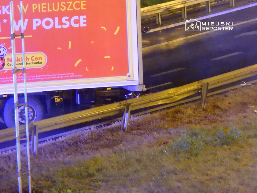 Tir z reklamą pieluszek stoi przy uszkodzonej barierce na poboczu drogi w nocy.
