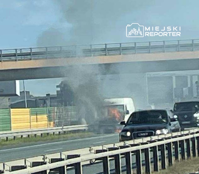 Dym i ogień wydobywają się z tyłu białego dostawczego samochodu na autostradzie, obok ruchu innych pojazdów.