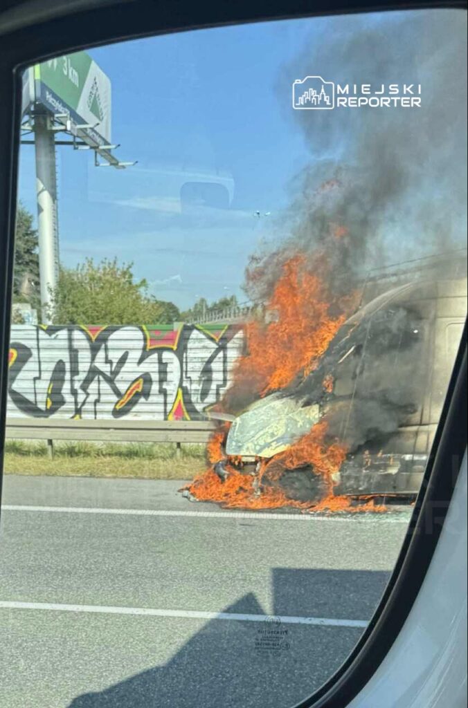 Van w płomieniach na poboczu drogi, obok graffiti na ogrodzeniu. Dym i ogień unoszą się w powietrzu.