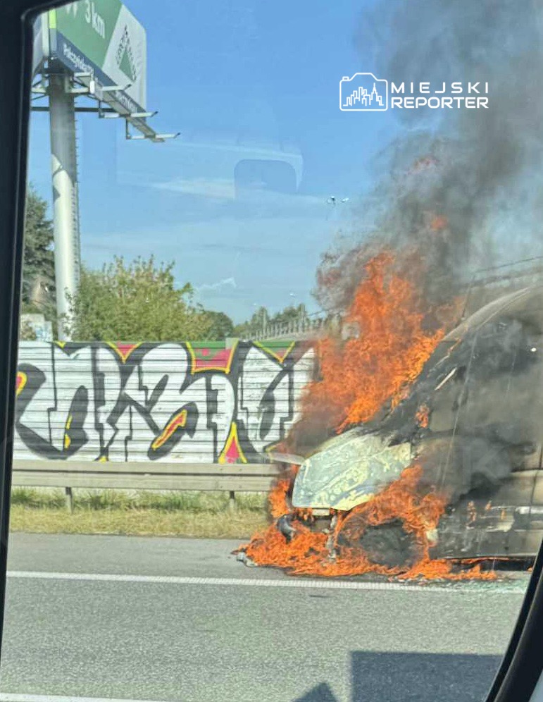Pojazd w płomieniach na pasie ruchu, obok graffiti na ogrodzeniu. Dym unosi się w niebo.