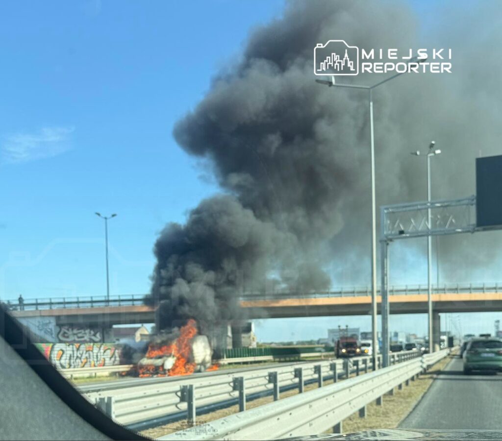 Na drodze autostrady samochód płonie, wznosząc czarne kłęby dymu, obok przejeżdżają inne pojazdy.
