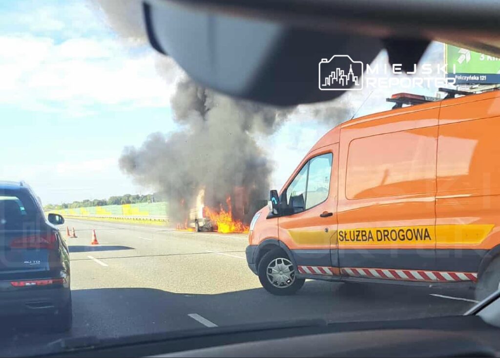 Na drodze pali się samochód, obok stoi pomarańczowy pojazd służby drogowej, a w powietrzu unosi się czarny dym.