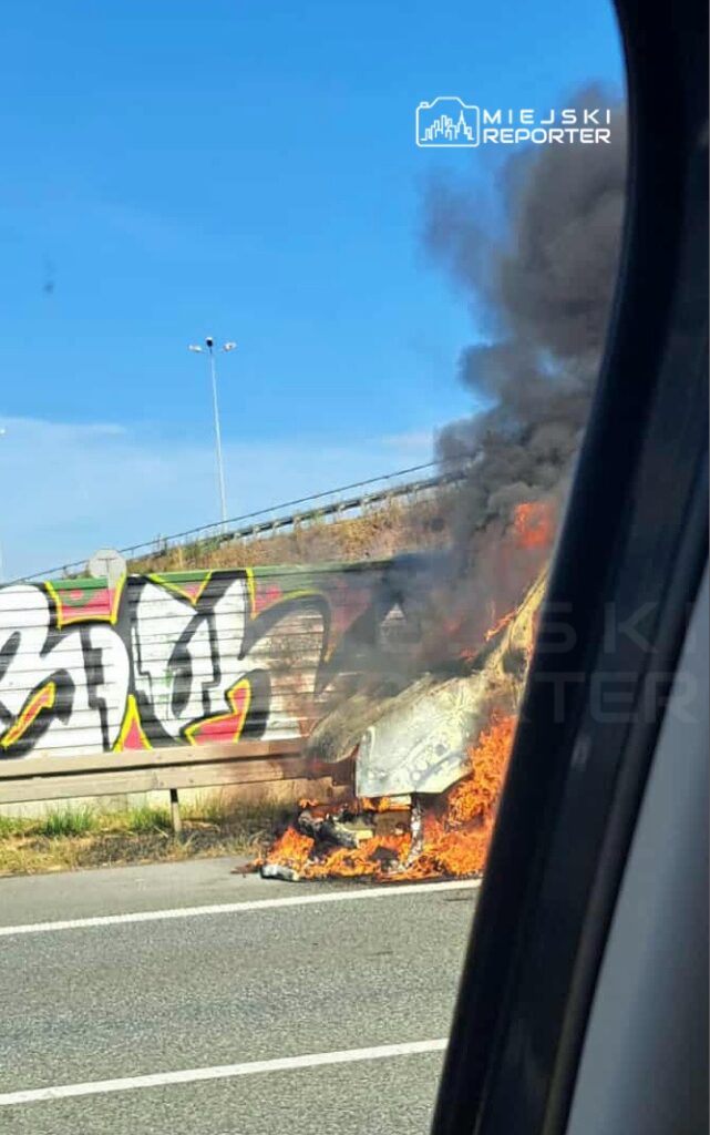 Pojazd płonący na poboczu drogi, wydobywający czarny dym, w tle graffiti na ścianie.
