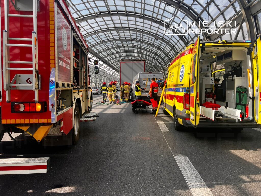 Zespół ratunkowy oraz strażacy udzielają pomocy poszkodowanemu na jezdni pod przeszklonym zadaszeniem.