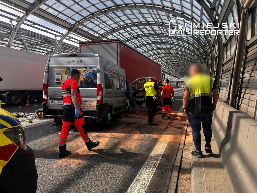 Ratownicy medyczni i policjanci pracują na miejscu wypadku w podziemnym tunelu, obok uszkodzonego pojazdu dostawczego.