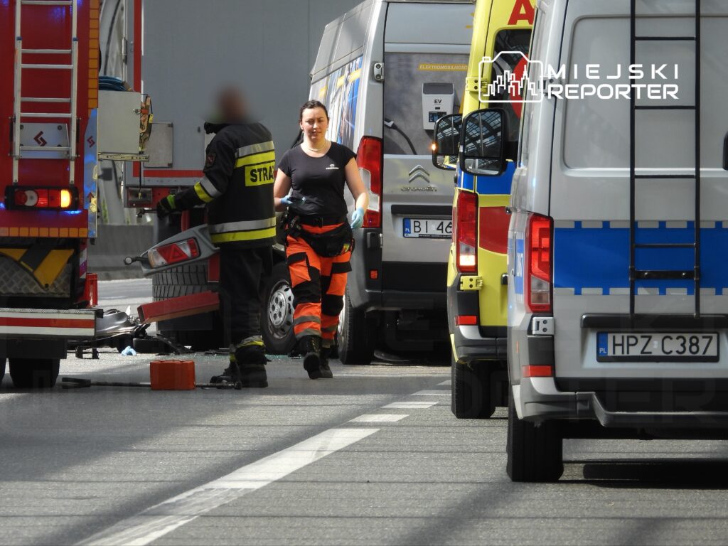Kobieta w mundurze strażaka idzie w kierunku pojazdów ratunkowych na drodze, obok leżą fragmenty szkła.