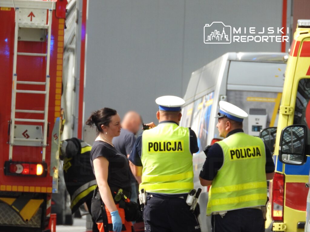 Kobieta w rękawiczkach rozmawia z dwoma policjantami w kamizelkach odblaskowych przy wozie strażackim.