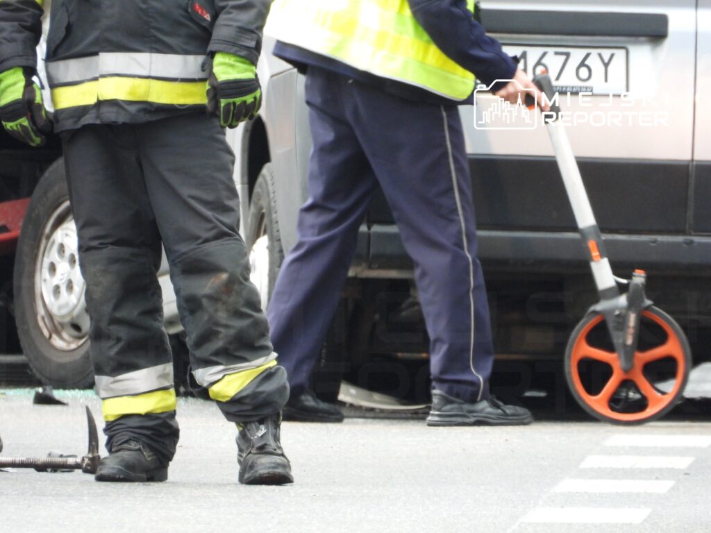 Strażak w mundurze z odblaskami mierzy odległość od uszkodzonego pojazdu na ulicy, obok leży sprzęt ratunkowy.