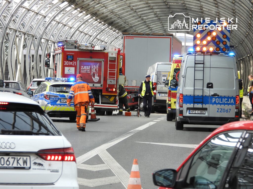 Na drodze zatrzymane pojazdy, w tym radiowozy i wóz strażacki, a pracownicy służb ratunkowych udzielają pomocy.