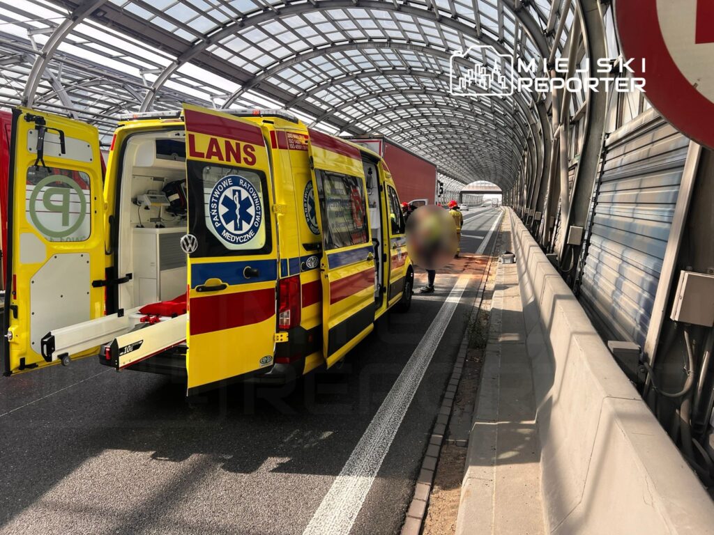 Zespół ratunkowy z otwartą tylną częścią karetki, przygotowujący się do akcji w nowoczesnym tunelu drogowym.
