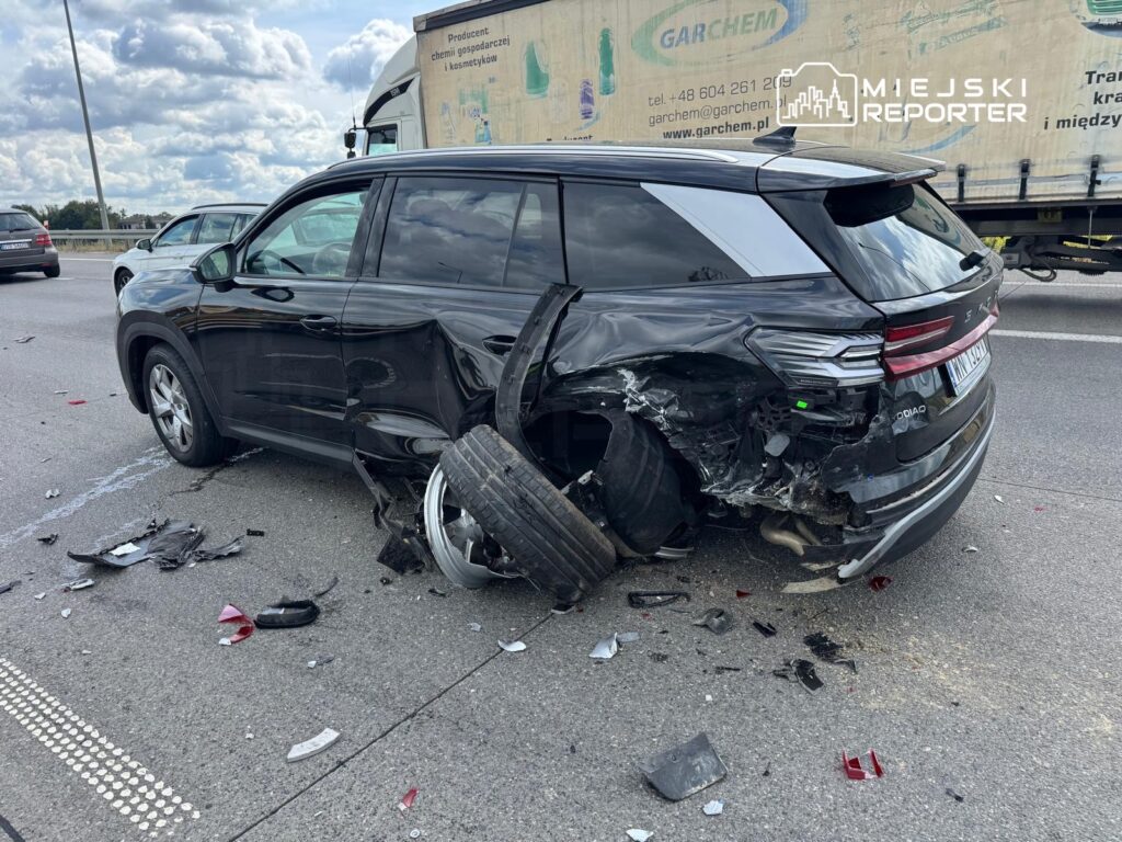 Czarny SUV z uszkodzoną tylną częścią stoi na drodze, wokół leżą resztki zderzaków i fragmenty opon.