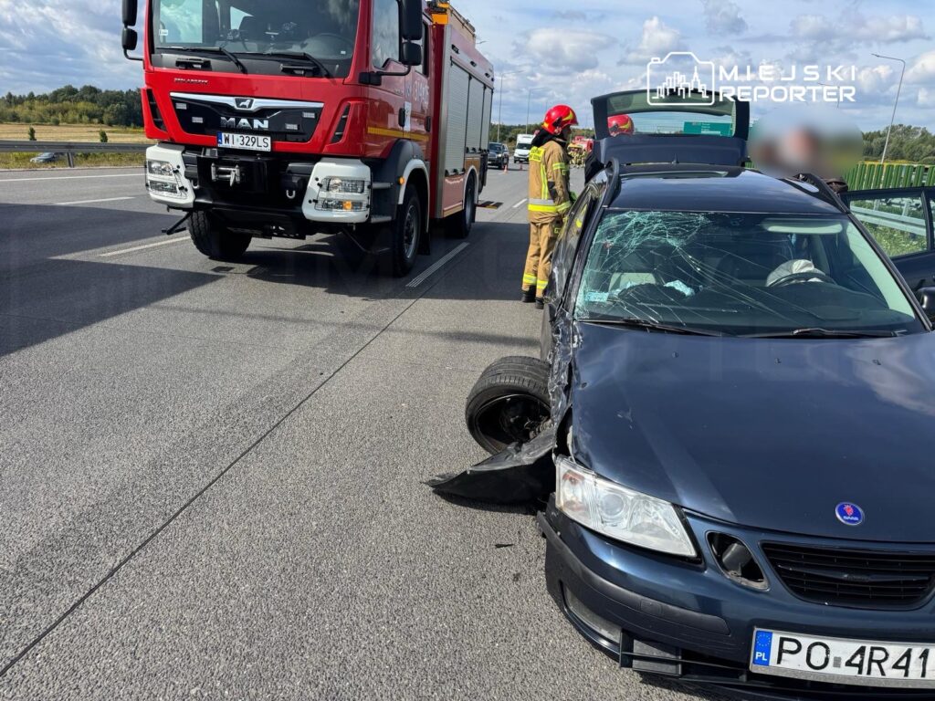 Czarny samochód osobowy z uszkodzoną przednią częścią stoi na drodze, obok niego strażacy w kaskach.