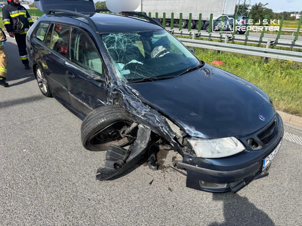 Uszkodzony samochód osobowy z oderwaną oponą i wgniecioną karoserią stoi na pasie drogi, obok grupy ratowników.