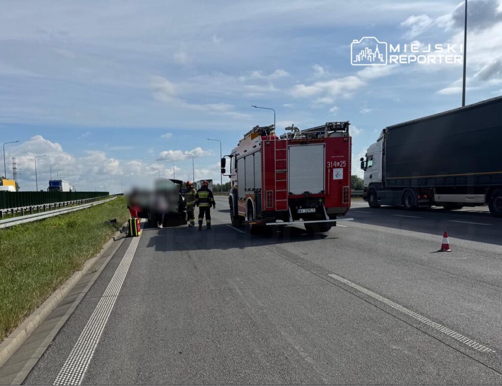 Strażacy udzielają pomocy poszkodowanemu w wypadku drogowym na autostradzie, obok zaparkowanego wozu strażackiego.