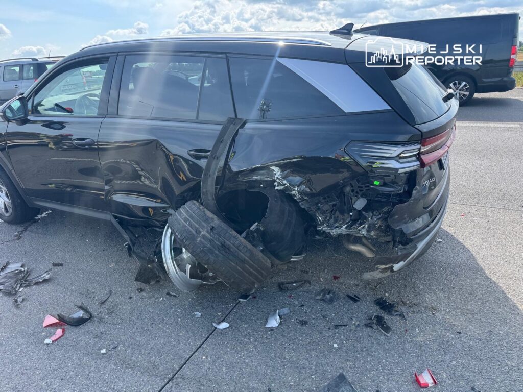 Czarny SUV z uszkodzoną tylną częścią, leżące kawałki plastiku i opona na drodze. W tle inne pojazdy na parkingu.