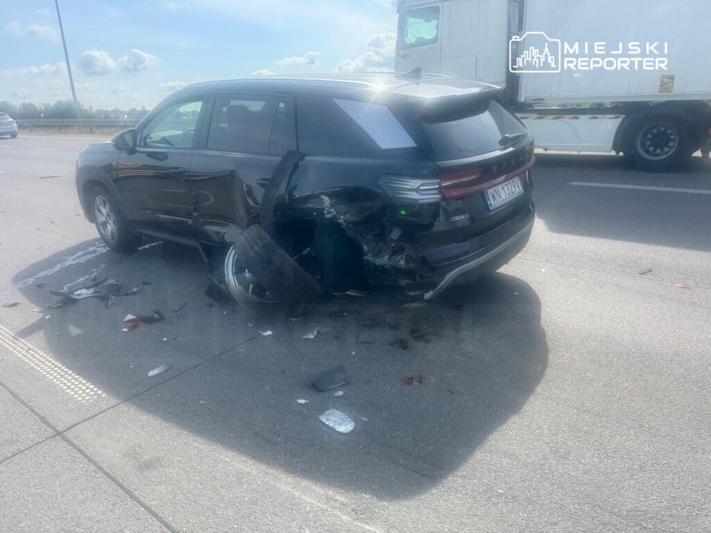 Czarny SUV z uszkodzoną tylną częścią stoi na drodze, obok leżą fragmenty zderzaków i opon.