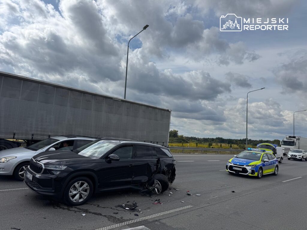 Czarne SUV z uszkodzoną tylną częścią stoi na autostradzie, obok radiowóz policji oraz inne pojazdy.
