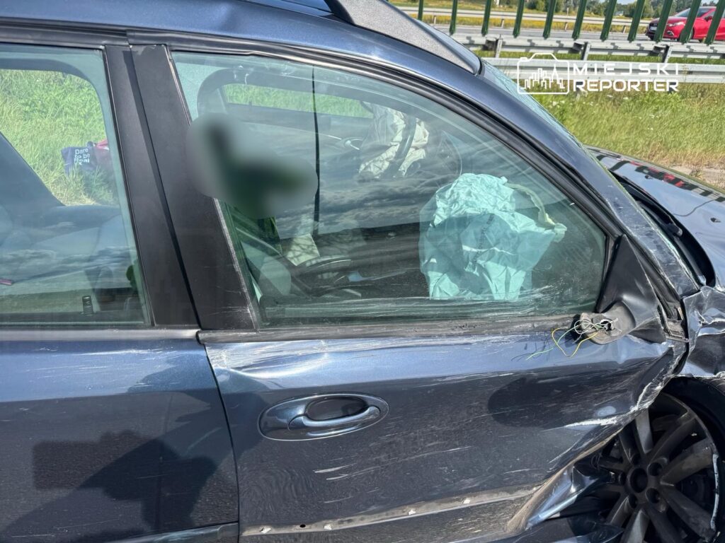 Uszkodzony samochód osobowy z wgniecioną stroną i wystrzeloną poduszką powietrzną, zaparkowany na poboczu drogi.