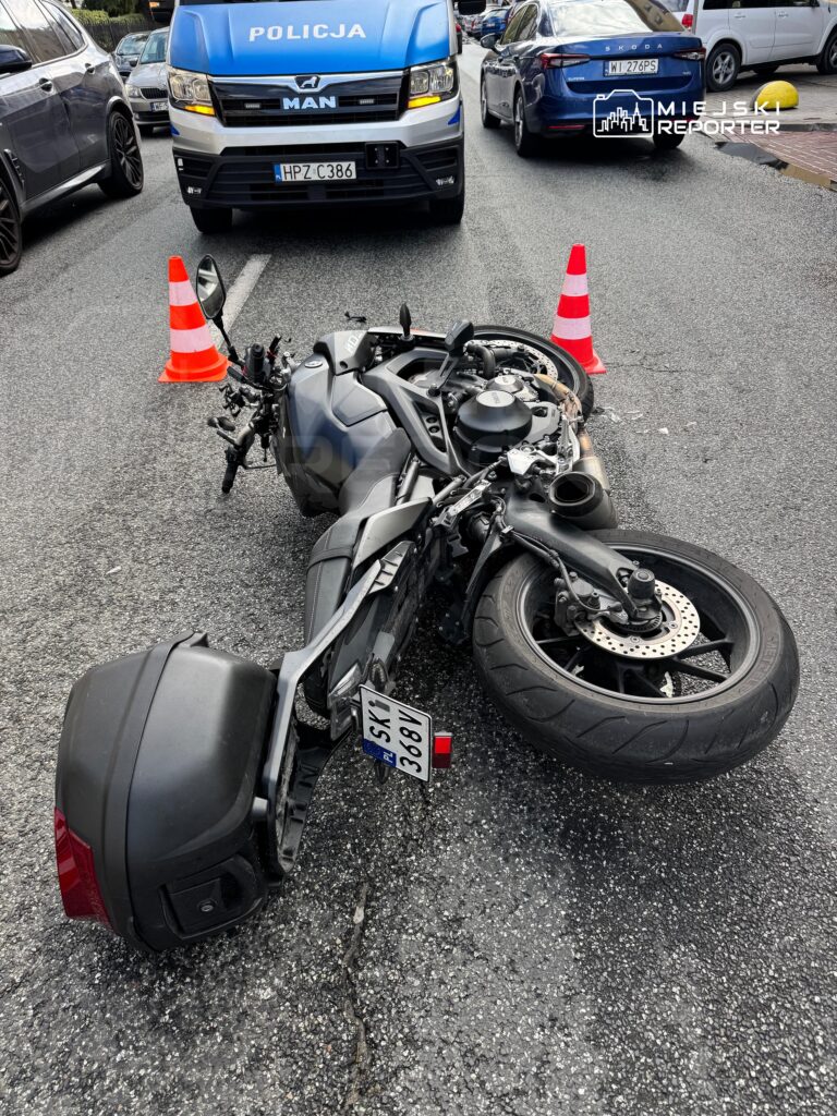 Motocykl leżący na jezdni, otoczony pachołkami drogowymi, obok radiowóz policyjny.