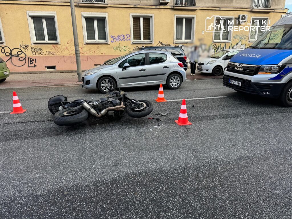 Motocykl leżący na jezdni, otoczony przez pachołki, obok policyjny radiowóz i zaparkowane samochody.