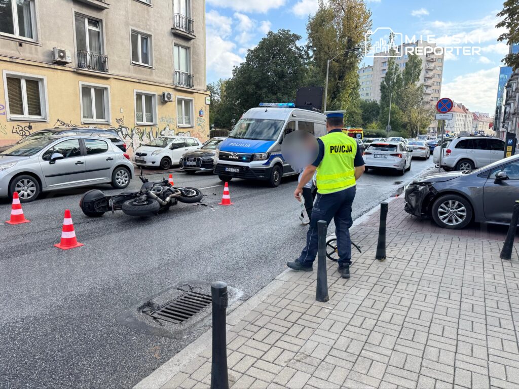 Motocykl leży na jezdni, otoczony pachołkami, obok policjant rozmawia z kierowcą uszkodzonego auta w miejskim otoczeniu.