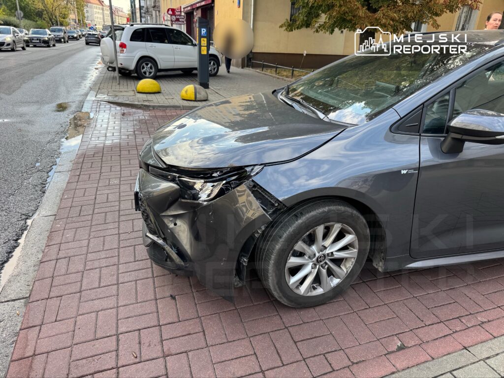 Uszkodzony samochód osobowy z wgniecionym przodem zaparkowany na chodniku obok ruchliwej ulicy.