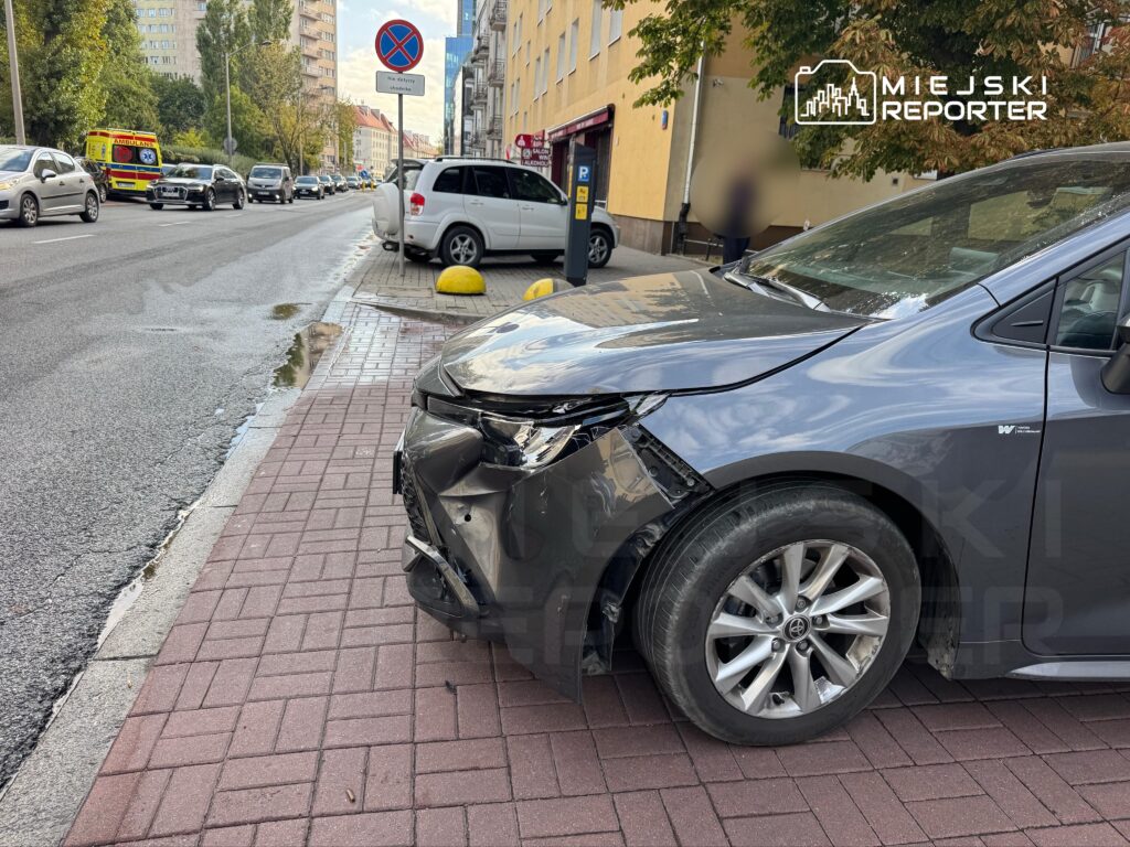 Uszkodzony samochód osobowy zaparkowany na chodniku, obok ruchliwej ulicy z przejeżdżającymi pojazdami.