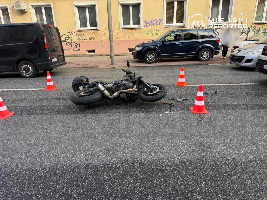 Motocykl leżący na drodze, otoczony przez pachołki drogowe, w pobliżu zaparkowanych samochodów.