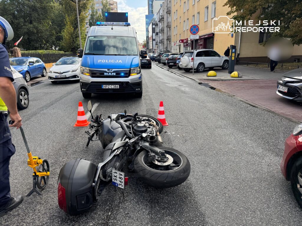 Motocykl leżący na jezdni, obok którego stoi policjant z urządzeniem pomiarowym, a w tle widać radiowóz.