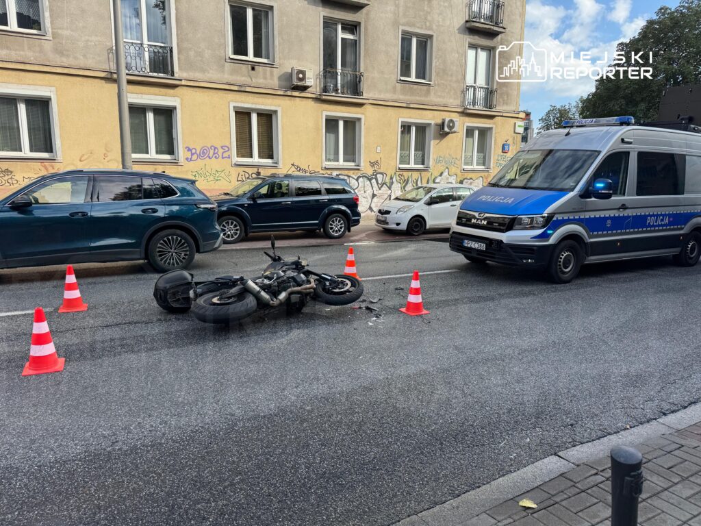 Motocykl leży na jezdni, otoczony pachołkami drogowymi, obok zaparkowane samochody oraz radiowóz policyjny.