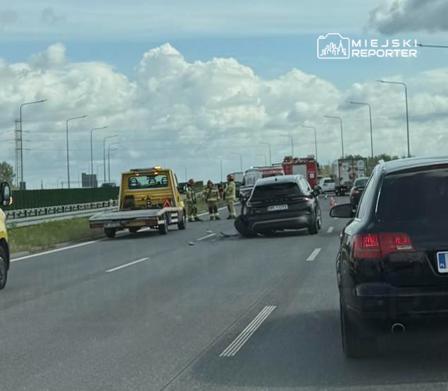 Na drodze stoją strażacy i ratownicy, obok uszkodzony samochód, a w tle widać pojazd pomocy drogowej.