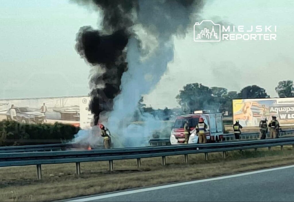 Strażacy gaszą pożar pojazdu na poboczu drogi, obok widoczna karetka i dym unoszący się w powietrzu.