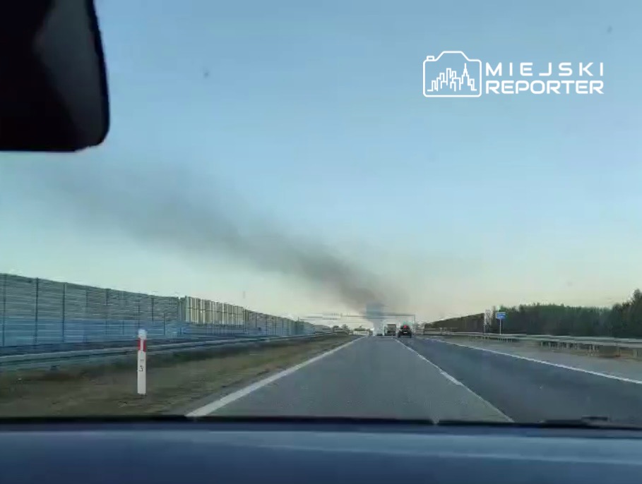 Samochód poruszający się po drodze, w tle widoczny dym unoszący się nad horyzontem.