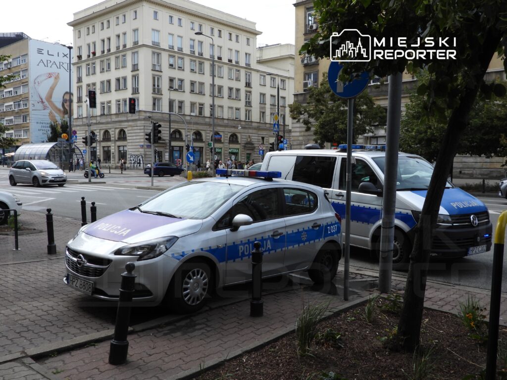 Policyjny samochód osobowy i furgonetka zaparkowane na chodniku w miejskim otoczeniu, obok ruchliwej ulicy.