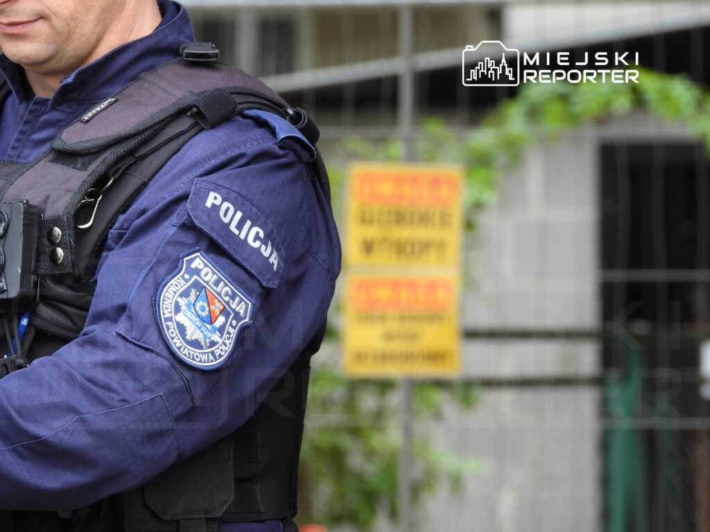 Policjant w mundurze z emblematem "Policja" stoi obok ogrodzenia z żółtymi tablicami ostrzegawczymi.