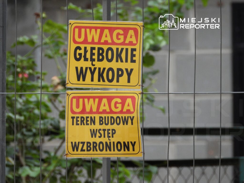Dwa żółte znaki ostrzegawcze informujące o głębokich wykopach oraz zakazie wstępu na teren budowy, umieszczone na ogrodzeniu.