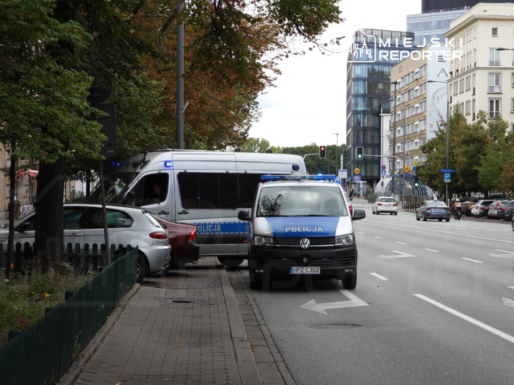Policyjny pojazd zaparkowany na ulicy, obok białego samochodu osobowego, w otoczeniu zieleni i miejskich budynków.