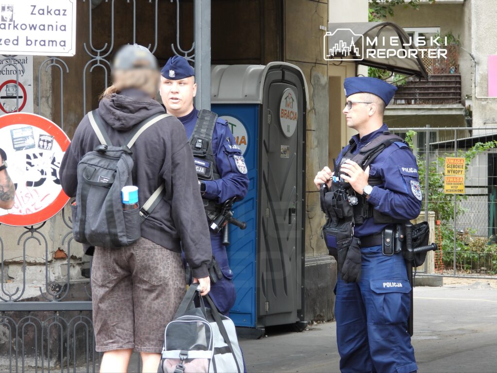 Dwaj policjanci rozmawiają z osobą w ciemnej odzieży na ulicy, w tle widoczny znak zakazu parkowania.