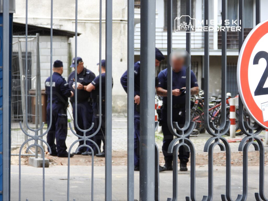 Grupa policjantów w mundurach rozmawia na podwórku, w tle stoją rowery i znak drogowy z ograniczeniem prędkości.