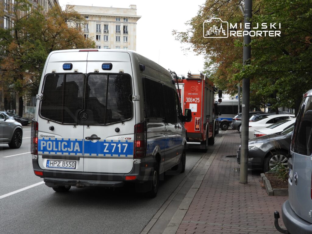 Policyjny furgon zaparkowany na ulicy, obok wozu strażackiego, otoczony samochodami osobowymi.