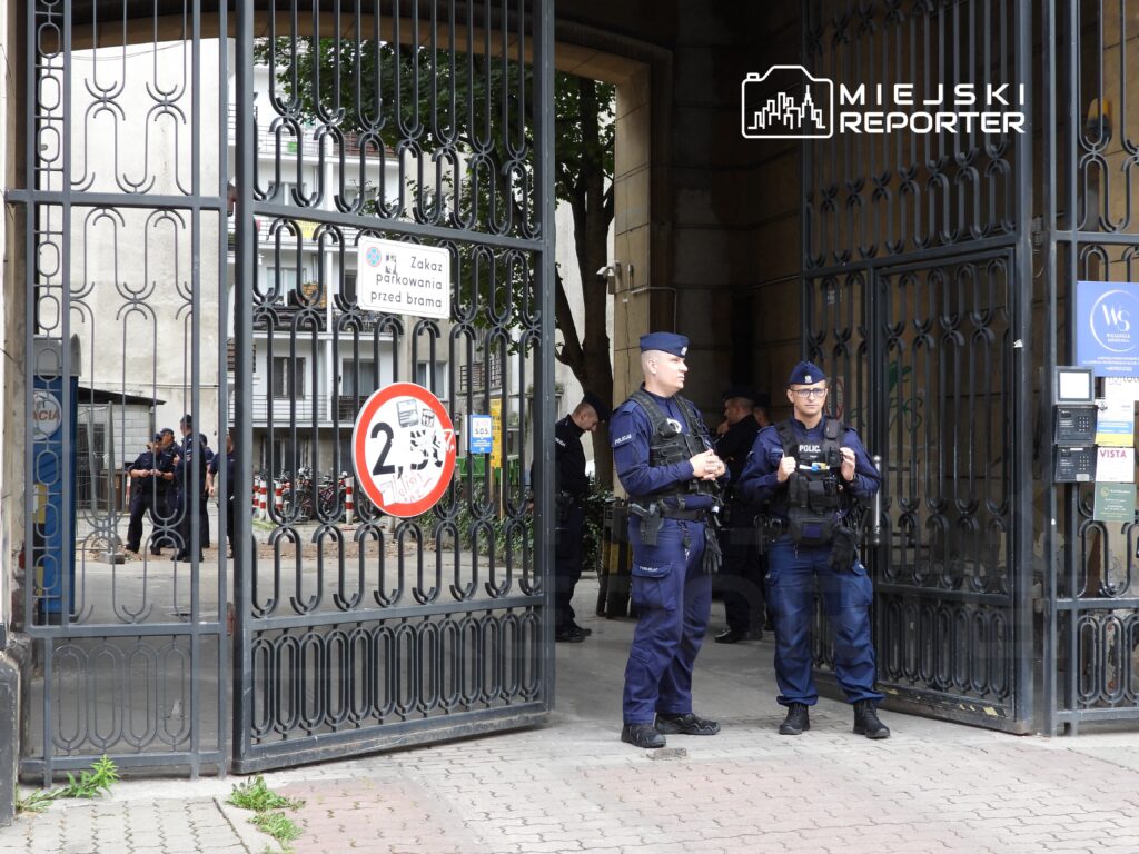 Dwaj policjanci w mundurach stoją przy otwartych bramach, obserwując otoczenie. W tle widać inne osoby oraz znaki drogowe.