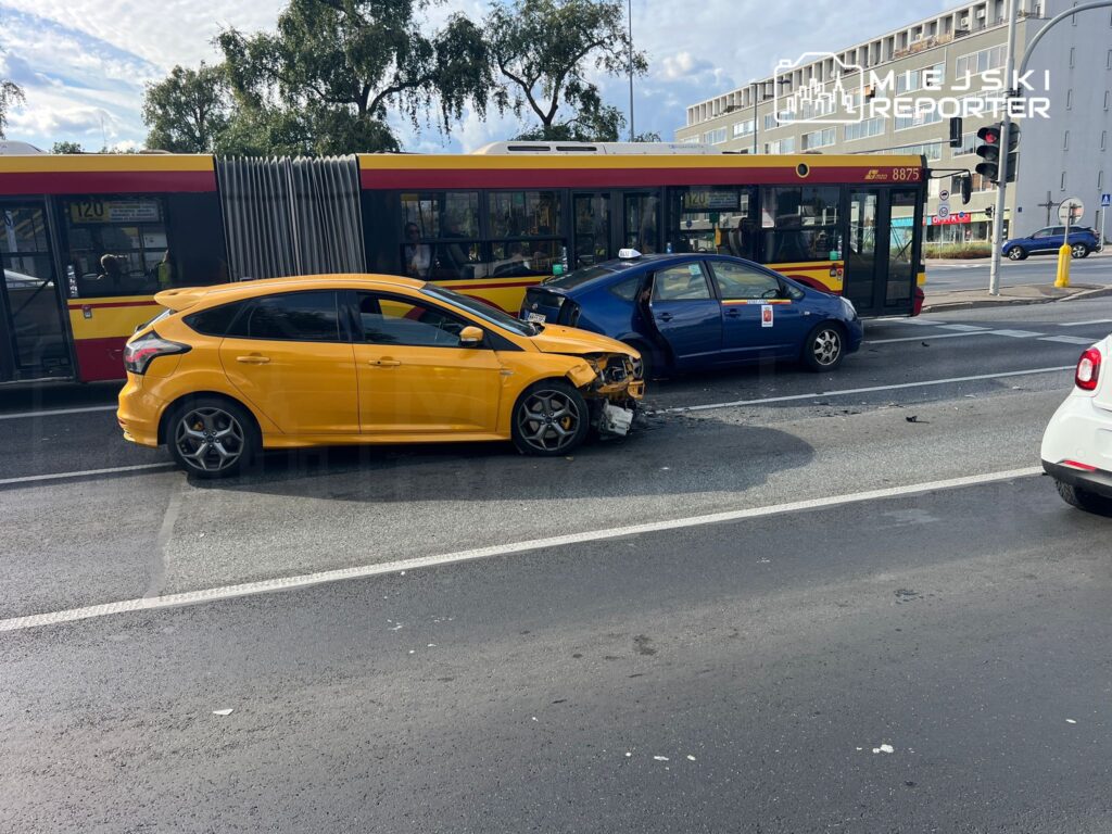 Zderzenie dwóch samochodów osobowych na ulicy, obok przystanku autobusowego. Żółty hatchback uszkodzony z przodu, niebieski sedan z otwartymi drzwiami.