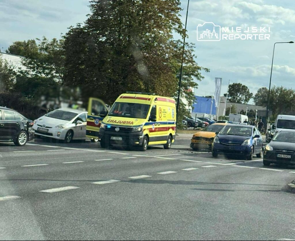 Karetka pogotowia z włączonymi sygnałami świetlnymi stoi w zatorze drogowym obok uszkodzonego samochodu.