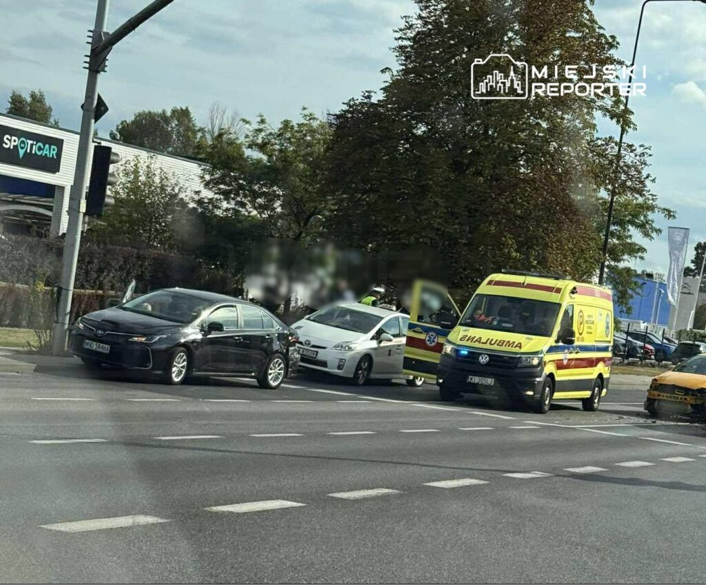 Czarny samochód osobowy stoi na skrzyżowaniu obok białego pojazdu, a obok nich zatrzymana karetka pogotowia.
