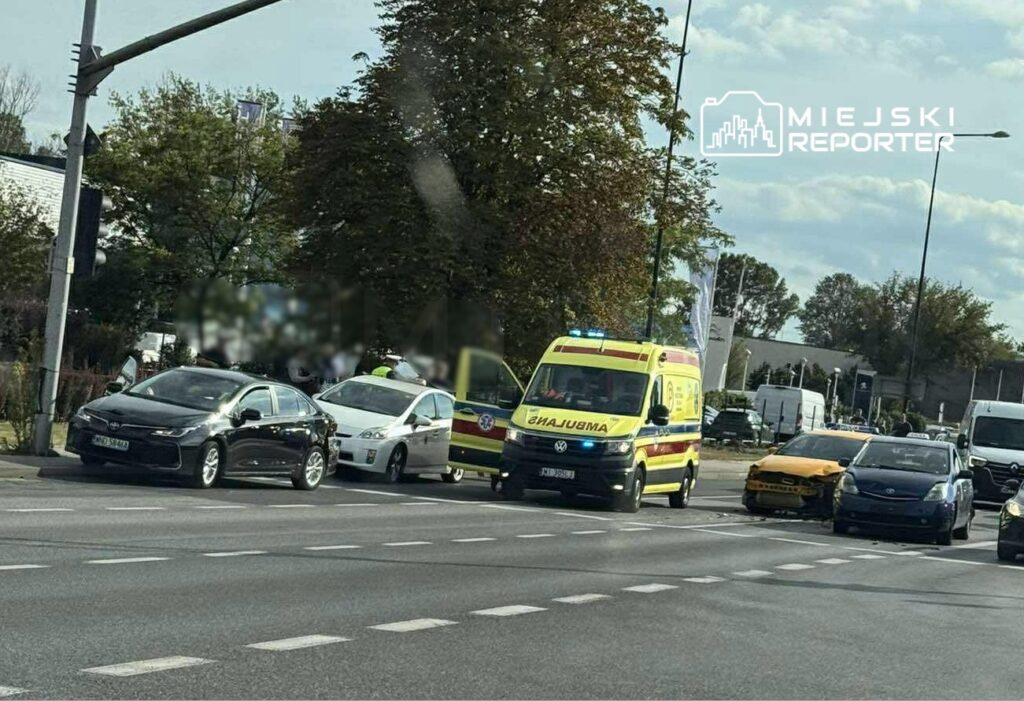 Na skrzyżowaniu stoją dwa uszkodzone samochody, obok nich znajduje się karetka pogotowia z włączonymi sygnałami.