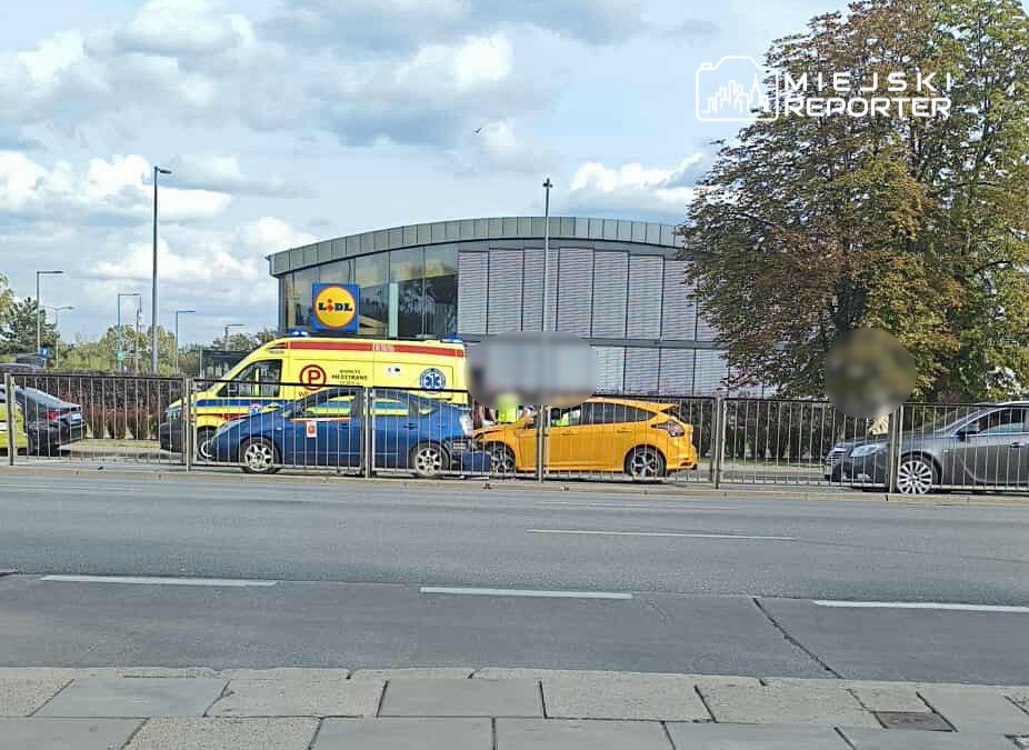 Karetka pogotowia ratunkowego zatrzymana obok parkingu sklepu, obok żółtego samochodu osobowego.
