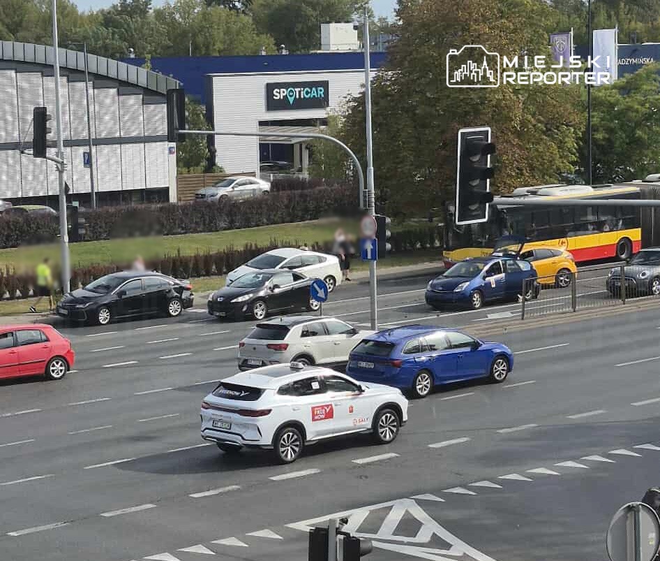 Ruchliwa ulica z kilkoma samochodami, w tym białym SUV-em, oraz przystanek autobusowy z żółtym autobusem.