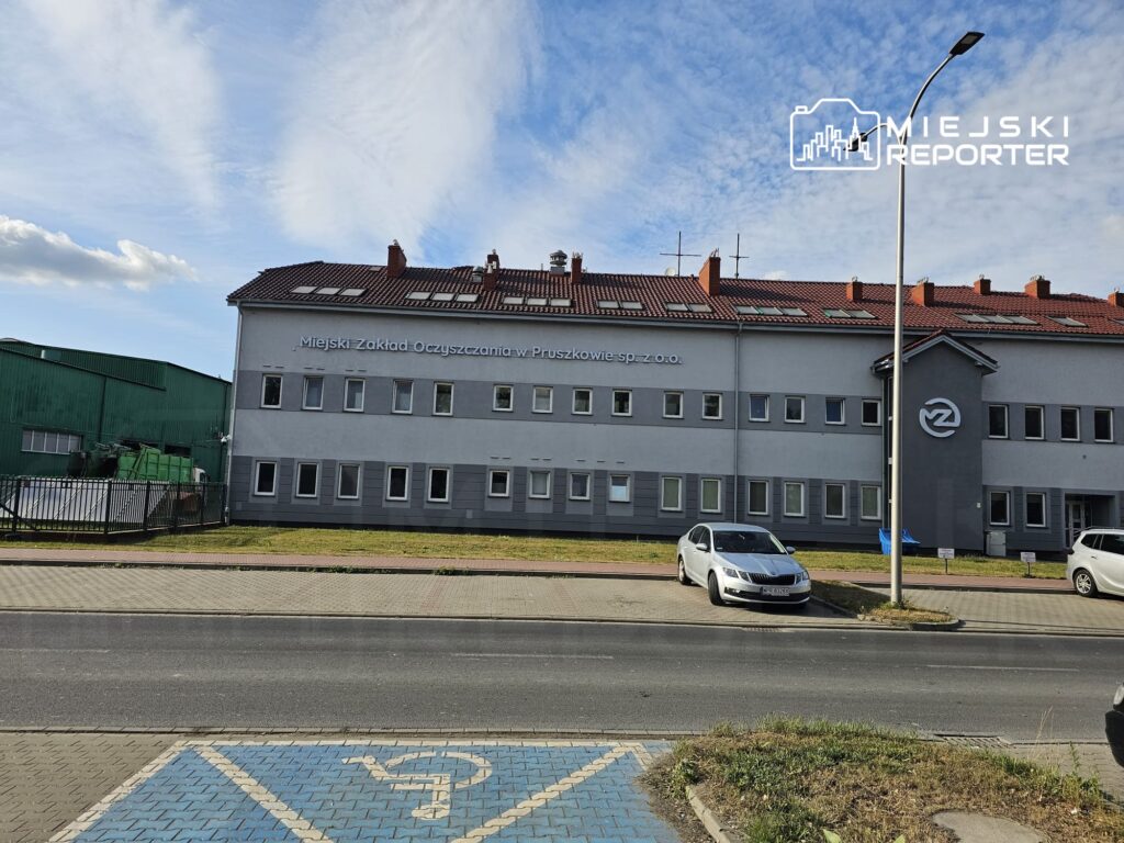 Miejski Zakład Oczyszczania w Pruszkowie, nowoczesny budynek z szaro-białą elewacją, otoczony zielenią i parkingiem.