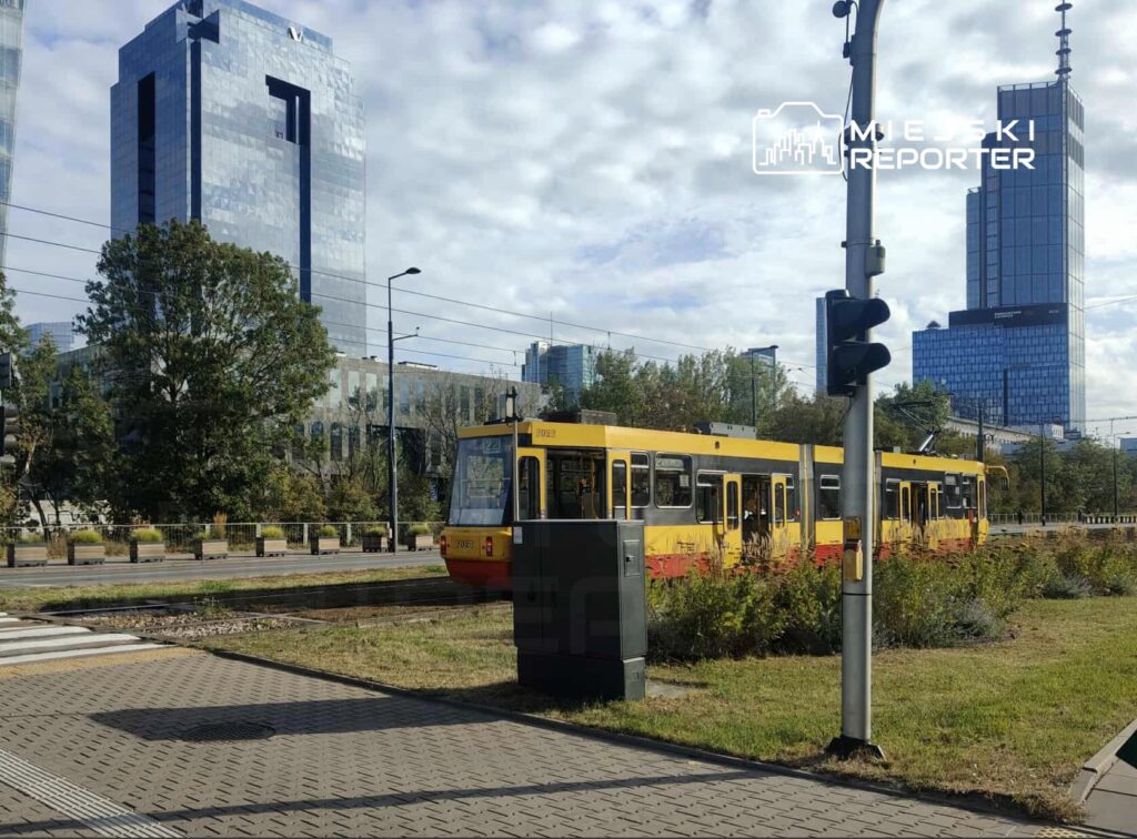 Żółty tramwaj zatrzymuje się na przystanku w nowoczesnej, miejskiej scenerii z wysokimi biurowcami w tle.