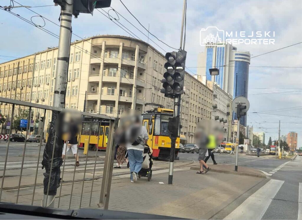 Grupa przechodniów przechodzi przez jezdnię obok tramwaju na przystanku w miejskim otoczeniu. W tle widać nowoczesne budynki.